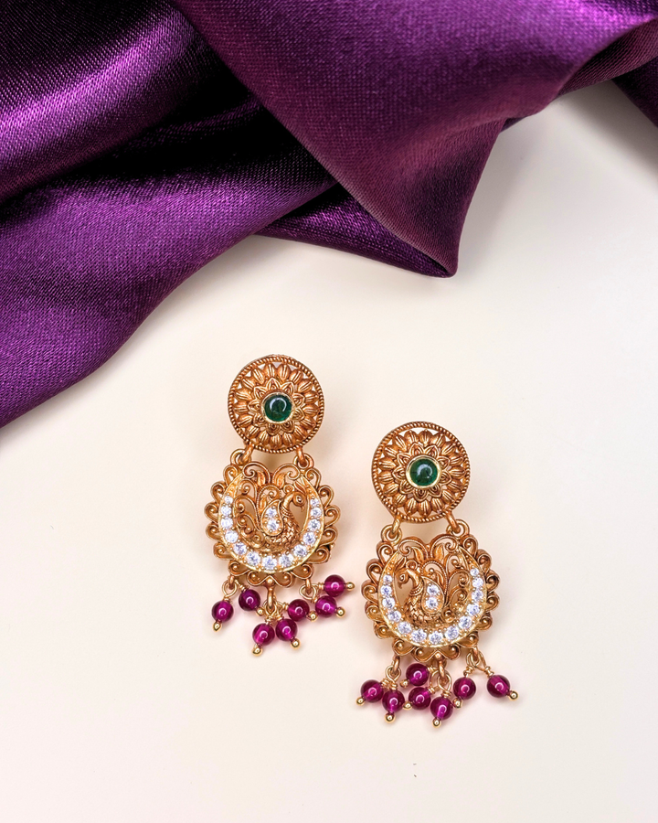 A pair of ornate gold-toned Indian jhumka earrings with green stone accents, white crystal details, and dangling magenta beads.