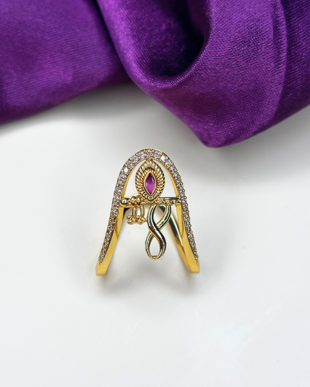A gold-toned ring designed in the shape of the letter 'A', embellished with clear crystals along the edges and a central teardrop-shaped pink gemstone