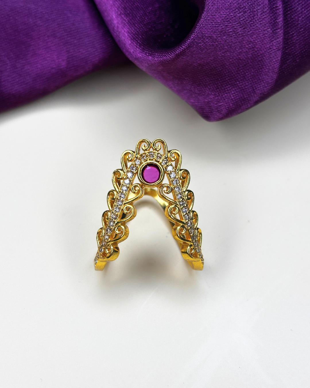 A gold U-shaped ring with intricate filigree detailing, clear pavé stones, and a central round pink gemstone.
